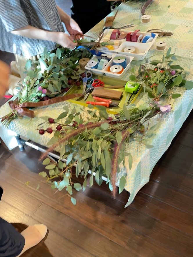 テーブルを囲んで、参加者それぞれがユーカリの枝を束ね、リボンで結んでいく様子。手前にはリボンの並んだトレー
